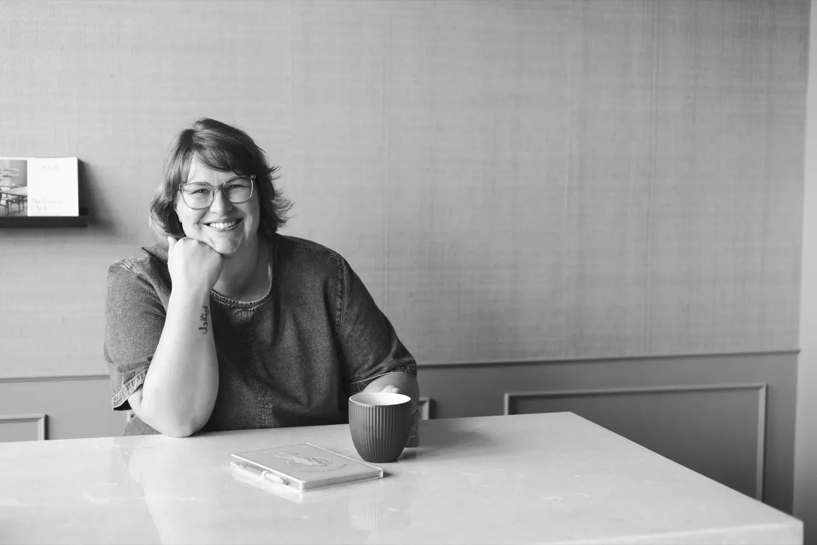 Black and white photo of Gemma sitting calmly at a table with a coffee and an open notebook, smiling.